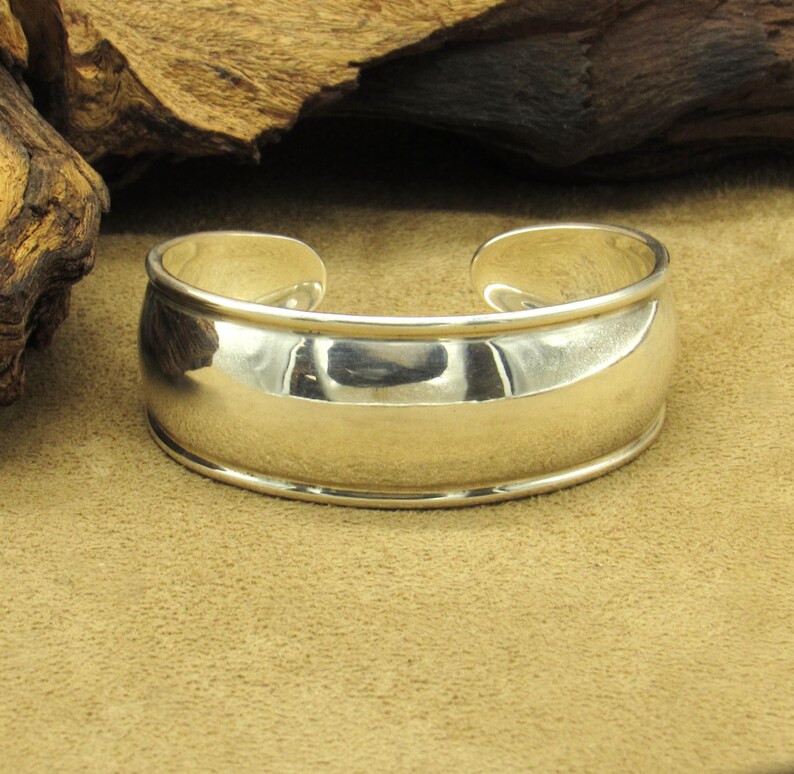 May include: A polished silver cuff bracelet. The bracelet has a wide, rounded band with a smooth, reflective surface. The design is simple and elegant, with a slightly curved shape that tapers towards the opening. The cuff is approximately 2.5 inches wide.
