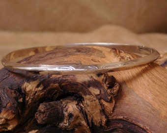 May include: A silver bangle bracelet with a hammered finish. The bracelet is on a wooden surface.