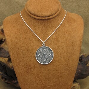 May include: A silver necklace with a round pendant featuring an intricate design of the Aztec calendar. The pendant is detailed and has a dark silver finish.