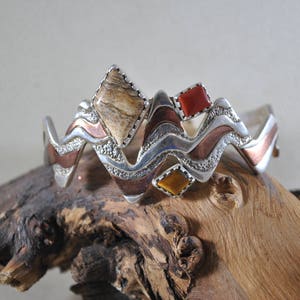 May include: A silver cuff bracelet with a wave-like design. It features inlaid stones in shades of brown, red, and yellow. The bracelet has a textured, layered appearance with copper and silver accents. The bracelet is displayed on a piece of driftwood.