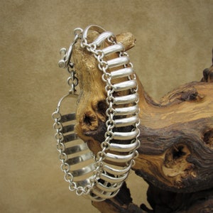 May include: A silver bracelet with a chain and a decorative, ridged band. The bracelet is displayed on a piece of driftwood.