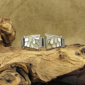 May include: A silver bracelet with a white mother-of-pearl inlay. The bracelet is made of two pieces that connect with a clasp. The bracelet is sitting on a piece of driftwood.