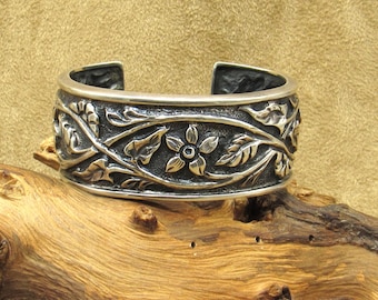 May include: A silver cuff bracelet with a floral and leaf design. The bracelet features a detailed pattern of flowers and foliage, with a dark background that highlights the design. The cuff is displayed on a piece of wood.