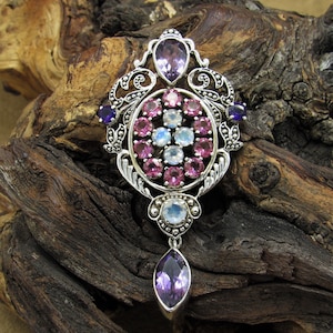 May include: A silver pendant with an ornate design, featuring a large teardrop-shaped amethyst at the bottom and a large amethyst at the top. The center has a circle of pink and blue gemstones.