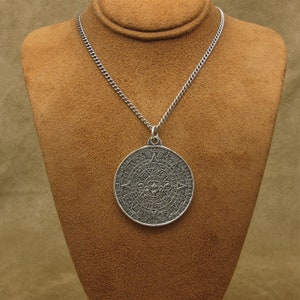 May include: A silver-colored pendant necklace featuring a large, circular pendant with an intricate Aztec calendar design. The pendant hangs from a delicate chain, displayed on a brown mannequin. The pendant's details are finely etched, showcasing the calendar's symbols.