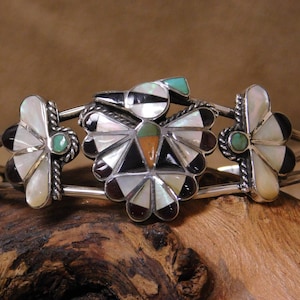 May include: A silver cuff bracelet with inlaid gemstones. The bracelet features a central design with a bird-like shape, surrounded by mother-of-pearl, black, and brown stones. Accented with turquoise and green stones, the bracelet has a Southwestern style.