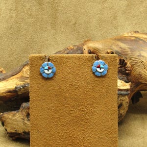 May include: A pair of flower-shaped earrings with a blue, white, and red design. The earrings are displayed on a brown suede-like surface. The earrings have a silver-colored border and are likely made of inlaid stones.