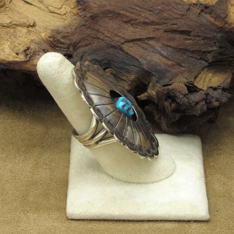 May include: A silver ring with a large turquoise stone set in a circular design. The ring is on a white display stand.