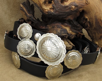 May include: A black leather belt with five silver concho belt buckles. The belt is folded and the buckles are visible.