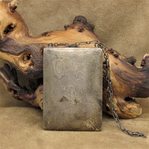 May include: A rectangular, hammered silver flask with a decorative chain. The flask features an engraved oval design within a shield shape. The background includes a piece of driftwood against a tan surface.