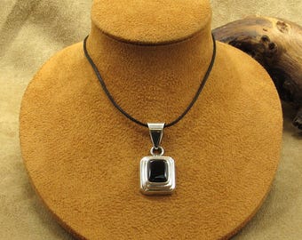 May include: A silver pendant necklace with a square black stone. The pendant is displayed on a brown bust. The necklace has a black cord. The pendant is a square shape with a black stone in the center.