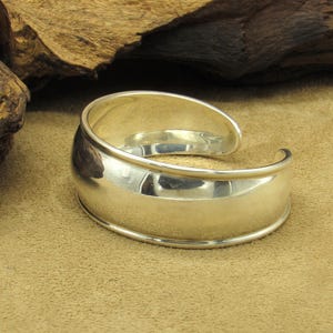 May include: A polished silver cuff bracelet with a curved design. The bracelet has a wide, flat center band and rounded edges. The open design allows for easy wear. The bracelet is displayed on a tan surface.