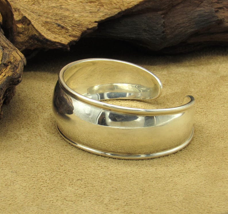 May include: A polished silver cuff bracelet with a curved design. The bracelet has a wide, flat center band and rounded edges. The open design allows for easy wear. The bracelet is displayed on a tan surface.