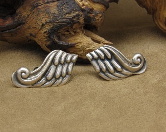 May include: A pair of silver earrings with a wing design and a swirl detail. The earrings are on a brown suede surface.