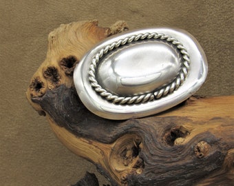 May include: A silver-toned oval belt buckle with a rope-like border. The buckle has a smooth, polished center and is set against a natural wood background. The buckle's design is simple yet elegant, suitable for various styles.