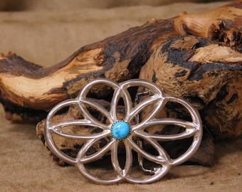 May include: A silver belt buckle with a turquoise stone in the center. The buckle is shaped like a flower with a geometric design.