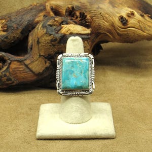 May include: A silver ring with a large, square turquoise stone. The turquoise is a vibrant blue-green color, set in a silver frame with a detailed rope-like border. The ring is displayed on a white ring holder against a neutral background.