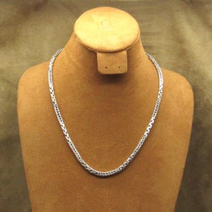 May include: A silver-colored chain necklace with a woven design is displayed on a brown jewelry bust. The necklace is made of small, interconnected links, creating a textured appearance. The bust is set against a neutral background.