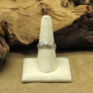 May include: A silver ring with an intricate floral design. The ring is displayed on a white ring holder against a natural wood background. The ring's detailed pattern is visible.