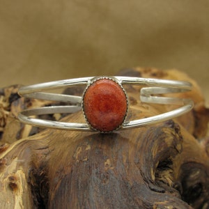 May include: A silver bracelet with a red coral stone set in the center. The bracelet is made of two thin bands of silver.