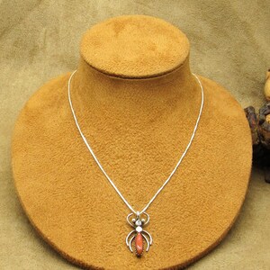 May include: A silver necklace with a pendant shaped like a stylized insect. The pendant features a central oval-shaped coral stone, surrounded by silver details. The necklace is displayed on a brown mannequin.