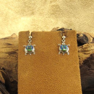 May include: A pair of silver-toned turtle earrings. Each earring features a small turtle pendant with blue, green, and silver accents. The earrings are displayed on a brown suede-like surface against a neutral background.