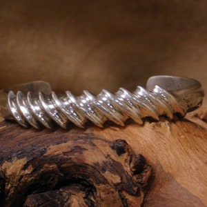 May include: A silver-toned cuff bracelet with a ridged, spiral design. The bracelet is displayed on a piece of dark, textured wood. The metal has a polished, reflective surface, and the overall aesthetic is modern and sculptural.