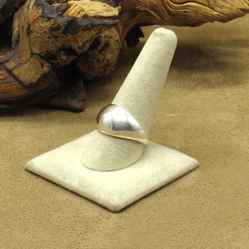 Peut inclure: Une bague en argent avec un dessus lisse et arrondi est pr&eacute;sent&eacute;e sur un pr&eacute;sentoir &agrave; bagues cr&egrave;me. La bague a une finition polie, refl&eacute;tant la lumi&egrave;re. C'est un bijou simple et &eacute;l&eacute;gant.