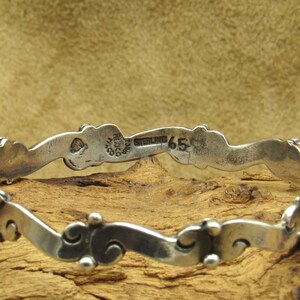 May include: A close-up of a silver bracelet with a wavy design. The bracelet has a series of decorative elements and small, round beads. The inscription "STERLING 65" is visible on the bracelet.