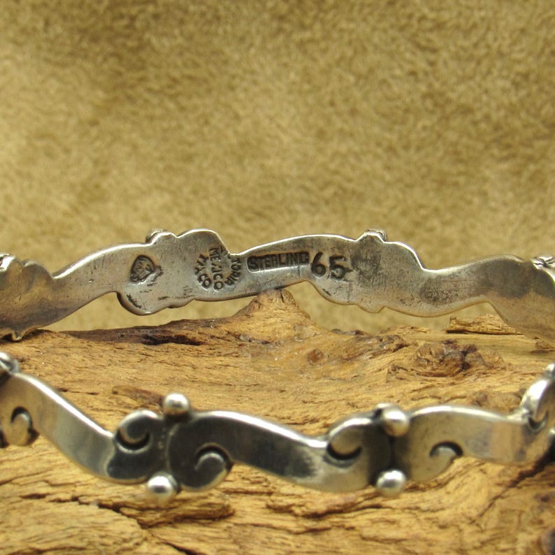 May include: A close-up of a silver bracelet with a wavy design. The bracelet has a series of decorative elements and small, round beads. The inscription "STERLING 65" is visible on the bracelet.