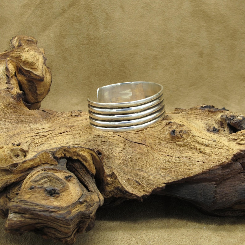May include: A silver cuff bracelet with five raised bands. The bracelet is on a wooden surface.