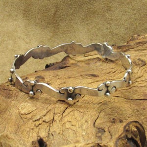 May include: A silver-toned bangle bracelet with a wavy design. The bracelet features a series of curved shapes and small, round embellishments. The bracelet is displayed on a piece of wood.