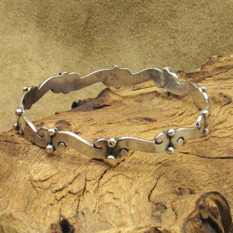 May include: A silver-toned bangle bracelet with a wavy design. The bracelet features a series of curved shapes and small, round embellishments. The bracelet is displayed on a piece of wood.