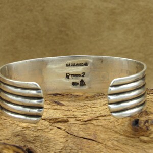 May include: Silver cuff bracelet with a ridged design. The back of the bracelet is stamped with "LA CUCHARACHA" and "RTKOO 2".