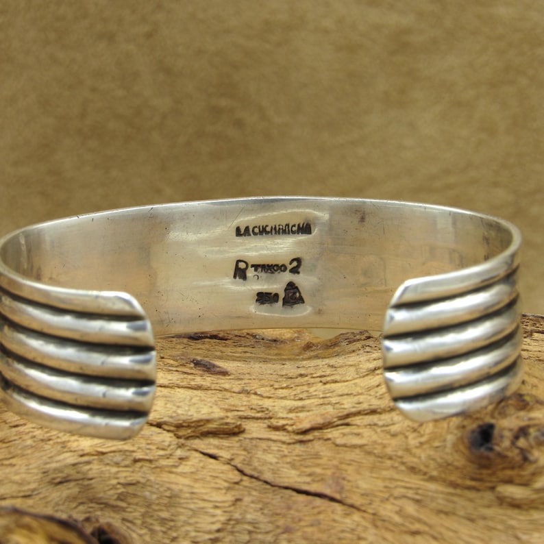 May include: Silver cuff bracelet with a ridged design. The back of the bracelet is stamped with "LA CUCHARACHA" and "RTKOO 2".