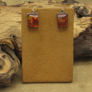 May include: A pair of square amber earrings with a silver-colored metal border. The earrings are displayed on a brown suede-like surface. The amber has a warm, orange-brown color.