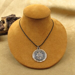 May include: A silver pendant necklace featuring a circular design with intricate details. The pendant hangs from a black cord and is displayed on a brown jewelry bust. The pendant has a dark center and a silver border.