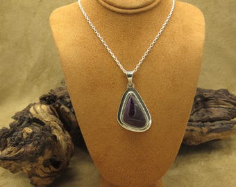 May include: A silver necklace with a pendant featuring a dark purple, triangular stone. The stone is set in a silver frame with a beaded edge. The necklace is displayed on a brown jewelry bust.