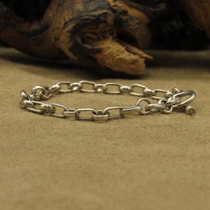May include: A silver chain bracelet with a simple, classic design. The bracelet features a series of interconnected oval links, secured with a circular clasp. The bracelet is displayed on a tan surface.
