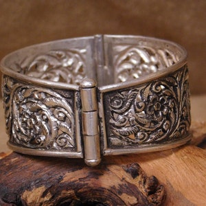 May include: A silver bracelet with an intricate floral design and a hinged clasp. The bracelet is made of silver and has a polished finish.