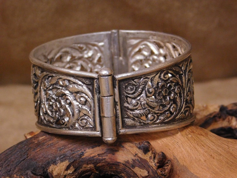 May include: A silver bracelet with an intricate floral design and a hinged clasp. The bracelet is made of silver and has a polished finish.
