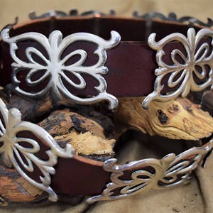 May include: A brown leather belt with silver butterfly-shaped buckles. The belt is wrapped around a piece of wood.