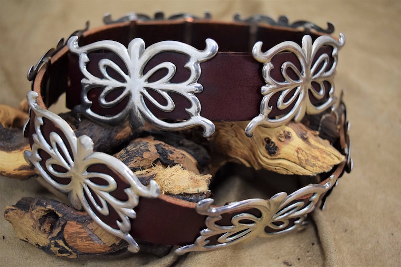 May include: A brown leather belt with silver butterfly-shaped buckles. The belt is wrapped around a piece of wood.