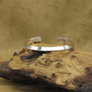 May include: A silver bangle bracelet with a simple, flat design. The bracelet is made of a single piece of metal that is bent into a circle.
