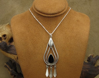 May include: A silver necklace with a black stone pendant. The pendant is shaped like a teardrop and has a delicate design. The necklace is on a brown velvet display stand.