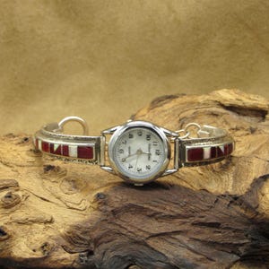 May include: A silver watch with a white face and black numbers. The watch band is made of silver and has red and white stones.