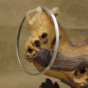 May include: A silver bangle bracelet with a decorative pattern around the edge. The bracelet is displayed on a piece of driftwood.