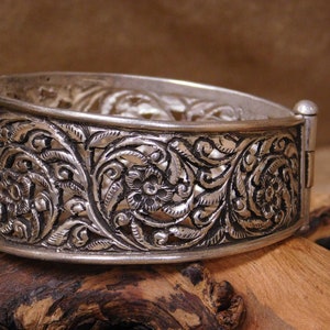 May include: A silver bangle bracelet with an intricate floral design. The bracelet is made of filigree and has a hinged clasp.
