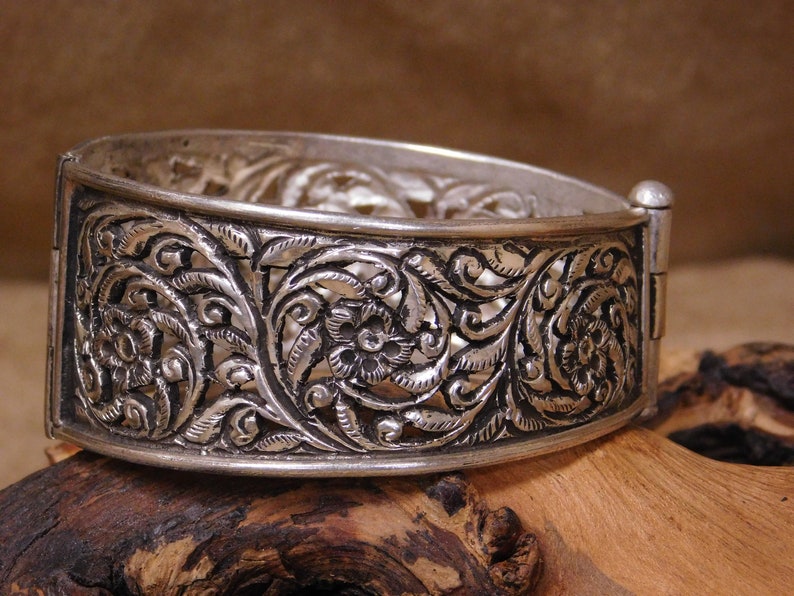 May include: A silver bangle bracelet with an intricate floral design. The bracelet is made of filigree and has a hinged clasp.