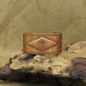 May include: A copper cuff bracelet with a geometric design. The bracelet is engraved with a diamond shape and a sunburst pattern.